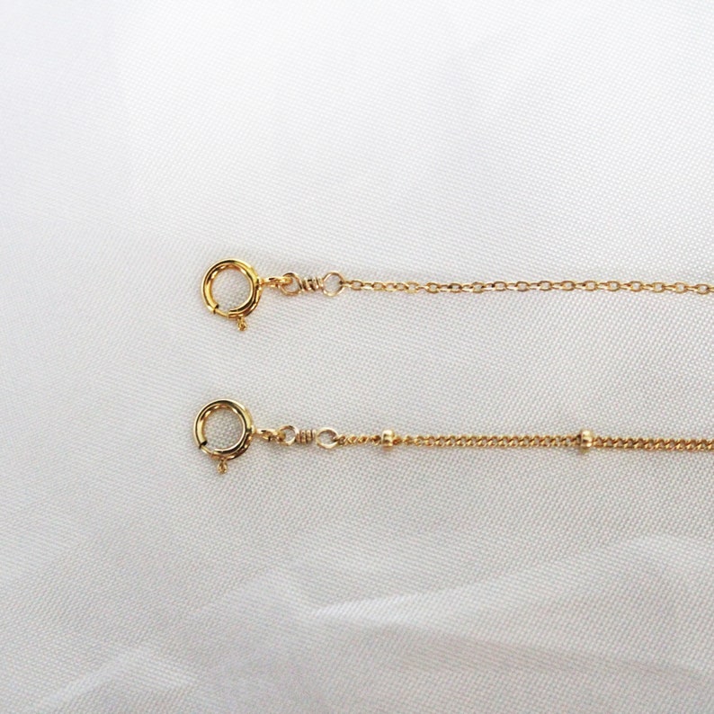May include: Two gold chains with lobster clasps. The top chain is a simple chain. The bottom chain has small gold beads.