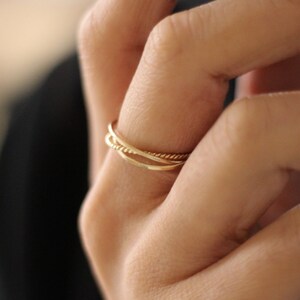 May include: A close-up of a hand wearing three gold rings. The rings are stacked on top of each other and are made of different styles. The ring on the bottom is a simple band, the middle ring is a twisted band, and the top ring is a textured band.