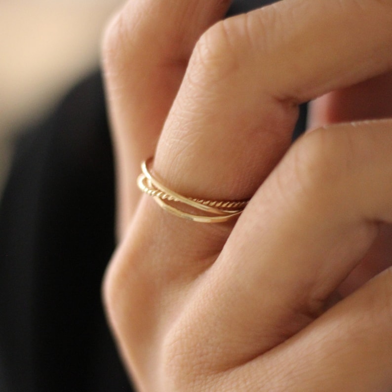 May include: A close-up of a hand wearing three gold rings. The rings are stacked on top of each other and are made of different styles. The ring on the bottom is a simple band, the middle ring is a twisted band, and the top ring is a textured band.