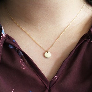 Puede incluir: Un collar de cadena dorada con un peque&ntilde;o colgante redondo. El collar lo lleva una persona con una blusa burdeos con estampado floral.
