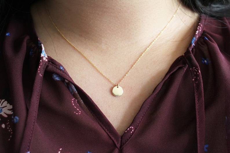 Puede incluir: Un collar de cadena dorada con un peque&ntilde;o colgante redondo. El collar lo lleva una persona con una blusa burdeos con estampado floral.