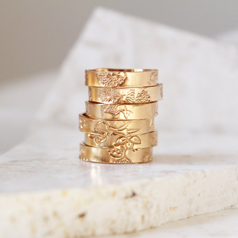 Puede incluir: Una pila de seis anillos dorados, cada uno con un dise&ntilde;o floral diferente grabado en la superficie. Los anillos est&aacute;n apilados uno encima del otro, mostrando los intrincados detalles de los patrones bot&aacute;nicos. Los anillos se exhiben sobre una superficie clara.