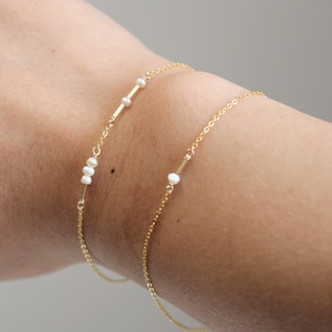 Peut inclure: Deux bracelets fins en or ornés de petites perles blanches. Les bracelets présentent un design de chaîne délicat, avec les perles regroupées en petits groupes. Les bracelets sont présentés sur un poignet, soulignant la simplicité élégante des bijoux.