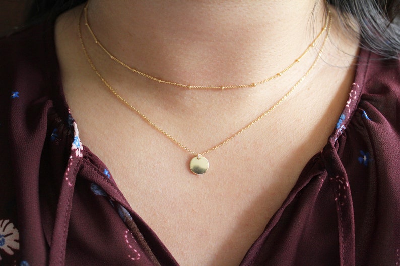 Puede incluir: Un collar de cadena de oro con un peque&ntilde;o colgante redondo. El collar est&aacute; superpuesto con una cadena m&aacute;s fina.