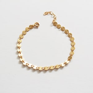 May include: A delicate gold bracelet featuring small, flat, circular charms linked together. The bracelet has an adjustable chain and a small clasp. The jewelry is displayed on a white surface.