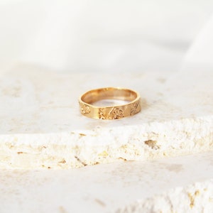 Puede incluir: Un anillo dorado con un dise&ntilde;o floral. El anillo est&aacute; sobre una superficie clara y texturizada. La banda presenta flores y hojas en relieve. El anillo es de estilo sencillo.