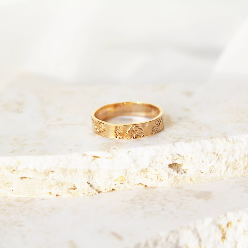 Puede incluir: Un anillo dorado con un dise&ntilde;o floral. El anillo est&aacute; sobre una superficie clara y texturizada. La banda presenta flores y hojas en relieve. El anillo es de estilo sencillo.
