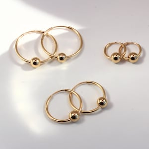 Op de afbeelding: Verschillende paren gouden oorringen met een kleine gouden bal accent. De oorbellen worden gepresenteerd op een witte ondergrond, met verschillende maten ringen. De oorbellen zijn gemaakt van een glanzend, reflecterend goud materiaal.