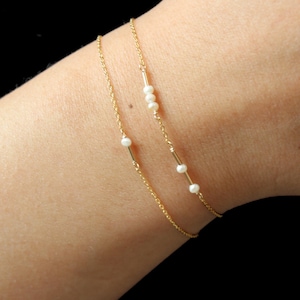 Peut inclure: Deux bracelets délicats en chaîne dorée. L'un comporte une perle unique, l'autre trois perles. Les bracelets sont portés sur un poignet sur fond noir. Les bracelets sont simples et élégants.