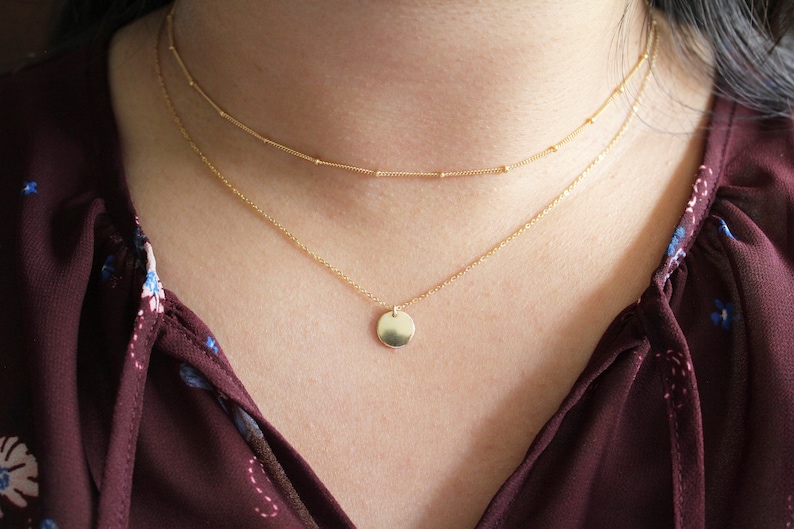 May include: A gold chain necklace with a small, round pendant. The necklace is layered with a thinner chain.