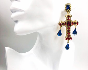 May include: A gold-tone cross earring featuring intricate filigree metalwork, embedded with multiple small red crystals, a central rectangular red stone, and three dangling blue teardrop-shaped crystals at the base and sides.