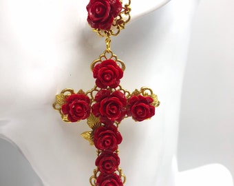 May include: A gold-tone drop earring featuring a cross shape adorned with multiple deep red rose blossoms. The design includes a single rose at the top post and a larger cross structure below, both framed by intricate gold-tone filigree metalwork.