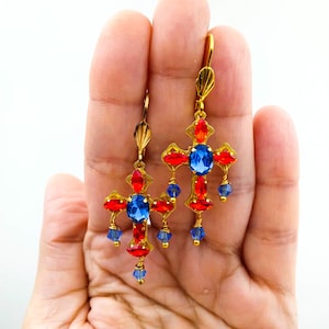 May include: Gold-toned cross earrings with red, blue, and gold accents. The earrings feature a cross design with a large blue center stone, red accents, and dangling blue beads. The earrings are held in a hand.