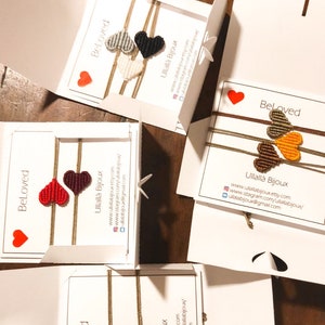 May include: Four white boxes with the word "Beloved" printed on them. Each box contains a set of friendship bracelets with different coloured heart designs. The bracelets are made of string and have a gold-coloured cord. The boxes also have the name "Ullallà Bijoux" and contact information printed on them.