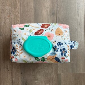 May include: A floral patterned baby wipes container with a teal lid that says "Pampers". The container has a blue bird, bees, and flowers on a white background. The container is rectangular and has a small fabric loop on the side.