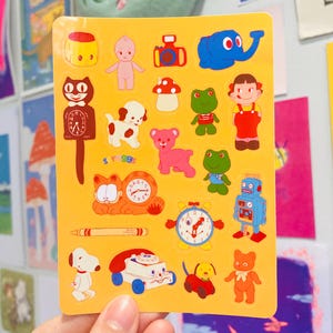 Puede incluir: Una hoja de pegatinas amarillas con varios personajes y objetos de dibujos animados. Las pegatinas incluyen un reloj, un robot, un perro, un gato, una rana, un oso, una cámara y un bebé. Las pegatinas son coloridas y tienen una estética vintage.