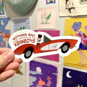 May include: A sticker featuring a red convertible car with white accents and a cartoon character. The text on the sticker reads "NOTHING BAD EVER HAPPENS TO THE KENNEDYS."