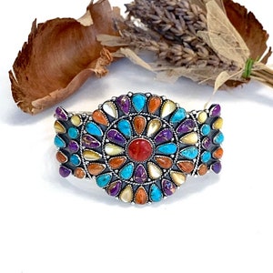 May include: A silver cuff bracelet with a floral design. The bracelet features a central red stone surrounded by turquoise, purple, orange, and white stones. The stones are set in a silver metal frame. The bracelet is displayed on a white surface.