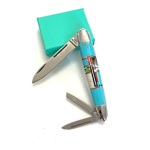 May include: A multi-tool pocket knife with a turquoise and inlaid design. The knife has a stainless steel blade and a decorative handle with a colourful geometric pattern. A small, teal-coloured box is in the background.
