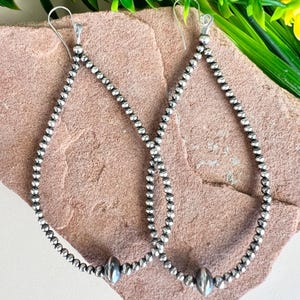 May include: A pair of silver teardrop earrings. Each earring is crafted from small, round beads strung together to form a teardrop shape. A larger, oval bead is centered at the bottom of each earring. The earrings hang from silver hooks.