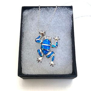 Puede incluir: Collar con colgante de rana plateada con incrustaciones de ópalo azul. La rana tiene las extremidades extendidas y se presenta en una caja de joyería negra con acolchado blanco. La cadena del collar es fina.
