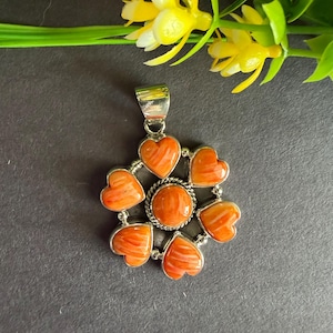 May include: A silver pendant featuring a central round orange stone surrounded by six heart-shaped orange stones. The stones have a marbled texture and are set in a polished silver frame with a twisted rope detail around the center stone.