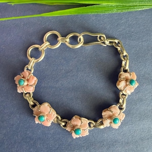 May include: A silver-toned metal chain bracelet featuring five carved light pink floral charms. Each flower is centred with a small, round, bright turquoise stone. The bracelet includes a series of circular links and a hook-style clasp closure.