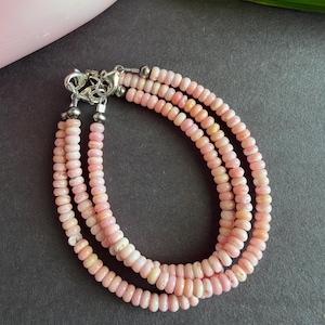Peut inclure: Un bracelet à trois rangs composé de petites perles roses rondes. Le bracelet est doté d'un fermoir argenté. Les perles sont d'un rose tendre, et le bracelet est probablement en pierre naturelle ou en verre.