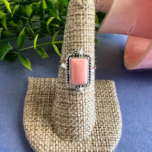 May include: A silver ring with a rectangular, light pink stone. The stone is set in a decorative silver frame. The ring is displayed on a tan ring holder. The ring is a piece of jewelry.