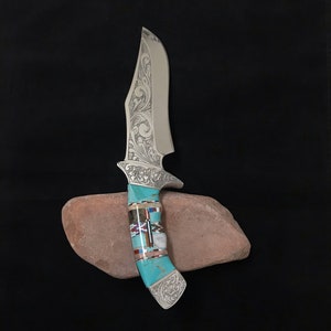 May include: A silver knife with a decorative handle made of turquoise and other stones. The knife is resting on a red rock.