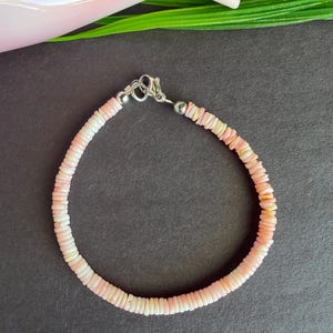 May include: A delicate bracelet composed of small, flat, pink and white shell beads. The bracelet is strung together with a silver-colored clasp. The beads are arranged in a circular pattern, creating a subtle, elegant accessory.