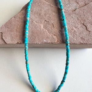 May include: A turquoise beaded necklace with a silver clasp. The necklace is made of small, round, turquoise-colored beads. The beads are strung together to form a necklace. The necklace is displayed on a brown surface.