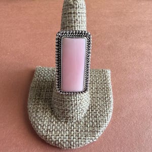 May include: A rectangular ring featuring a smooth, light pink stone set in a silver-toned metal frame. The setting is decorated with a beaded edge and a twisted rope-like border surrounding the central pink stone, creating a detailed design.