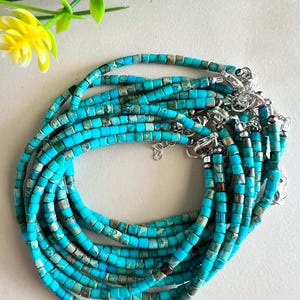 Puede incluir: Una colección de collares de cuentas turquesas con cierres plateados. Los collares están hechos de pequeñas cuentas cuadradas en tonos turquesa y gris. Los collares están dispuestos en un patrón circular.