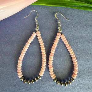 May include: A pair of teardrop-shaped earrings featuring light pink beads arranged in a loop, accented with five metallic silver beads at the bottom of each earring. The earrings are suspended from silver-toned metal fishhook ear wires.