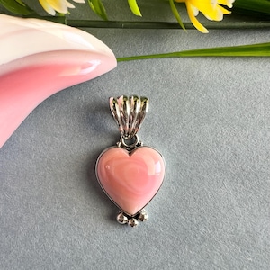May include: A heart-shaped pendant with a pink stone set in a silver-colored metal. The pendant has a bail at the top and small decorative balls at the bottom. The background is a light blue-gray color.