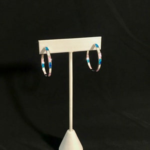 May include: A pair of silver hoop earrings with pink and blue opal inlay. The earrings are displayed on a white stand against a black background.