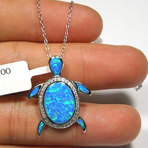 Puede incluir: Un collar de cadena de plata con un colgante de tortuga de ópalo azul. La tortuga está rodeada por una fila de piedras transparentes.