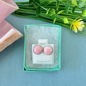 May include: A pair of pink stud earrings in a small, open, teal gift box. The earrings are round with a silver border. The box sits on a light blue surface, with greenery and a yellow flower in the background. The text "STERLING SILVER" is visible.