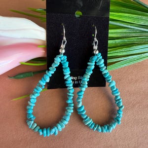 May include: A pair of teardrop-shaped dangle earrings featuring small, irregular, bright blue turquoise-colored stone chips strung on wire. The earrings are attached to a black rectangular display card with silver-toned metal hooks.