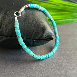 May include: Turquoise beaded bracelet with a silver clasp. The bracelet is made of small, round turquoise beads strung together. The clasp is silver and connects the two ends of the bracelet. The bracelet is displayed on a black surface.