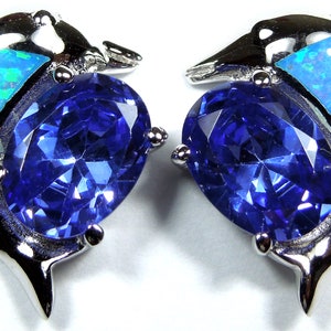 May include: A pair of silver dolphin earrings. Each earring has a large, oval, deep blue gemstone, with blue and green opal-like stones. The dolphins have a polished, reflective finish.