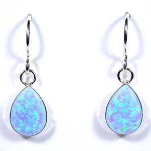 May include: A pair of silver hoop earrings featuring teardrop-shaped opal pendants. The opals display a light blue base with iridescent green and purple flashes. The earrings are set against a white backdrop.