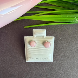 May include: A pair of round, light pink stud earrings set in sterling silver. The earrings are mounted on a white textured display card with the text STERLING SILVER embossed at the bottom, showcasing a simple and clean design for jewelry.
