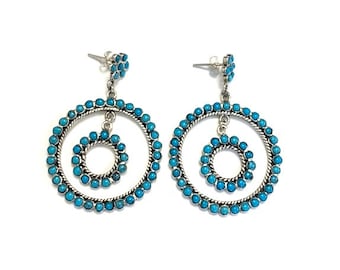 May include: A pair of silver dangle earrings with turquoise stones. The earrings feature two concentric circles, with the smaller circle hanging from the larger circle.