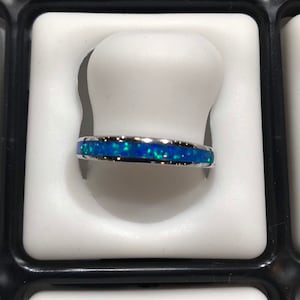 May include: A silver ring with a band of blue and green opal-like material. The ring is set in a white display case.