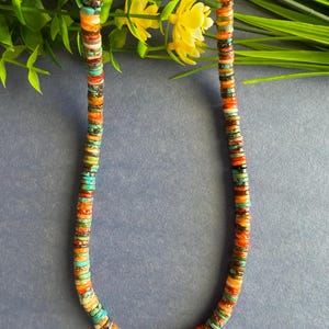 May include: A necklace composed of small, flat, disc-shaped beads in a vibrant mix of turquoise, orange, red, brown, and cream colors. The beads are strung together in a repeating pattern, creating a colorful, textured strand that hangs in a U-shape.