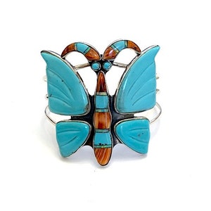 Può includere: Un bracciale a polsino in argento a forma di farfalla. Il design della farfalla presenta ali blu turchese e un corpo con accenti marroni e turchesi. Il bracciale è posto su uno sfondo bianco.