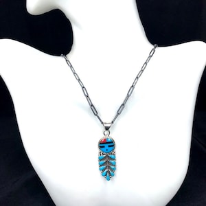 May include: A silver chain necklace with a turquoise and red pendant featuring a Native American design. The pendant is shaped like a feather with a face at the top.