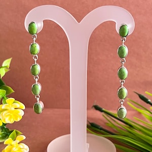 May include: A pair of long, dangling earrings with oval, light green stones set in silver. The earrings are displayed on a clear stand against a soft pink background, with yellow flowers and green foliage in the foreground.
